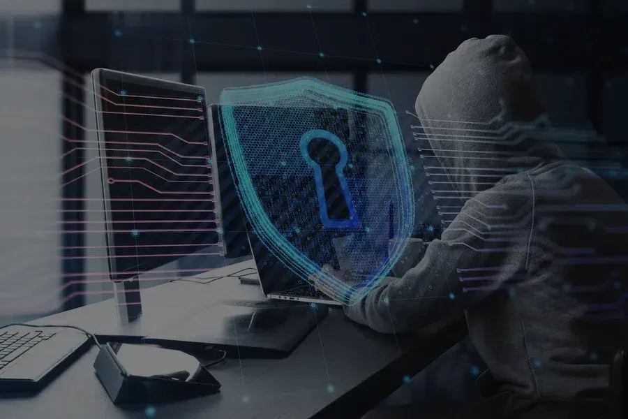 A hooded figure at a laptop behind a glowing padlock shield, the kind of attack a penetration test simulates